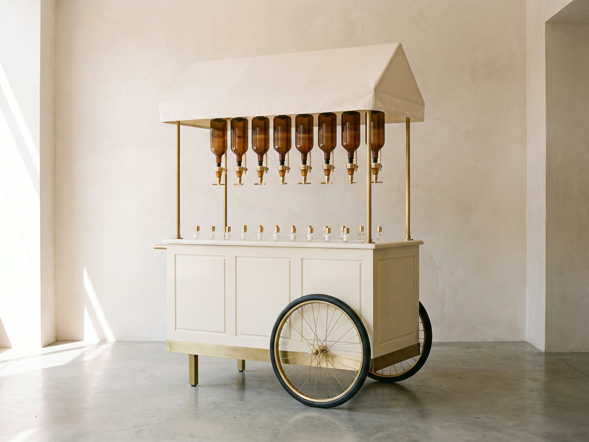 A mobile scent bar cart with a pitched cream canopy, eight amber dispensers on brass taps hanging beneath it