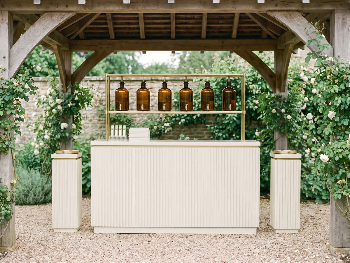 An outdoor scent bar with a fluted counter and flanking pedestals, amber dispensers on a brass frame beneath a garden pavilion