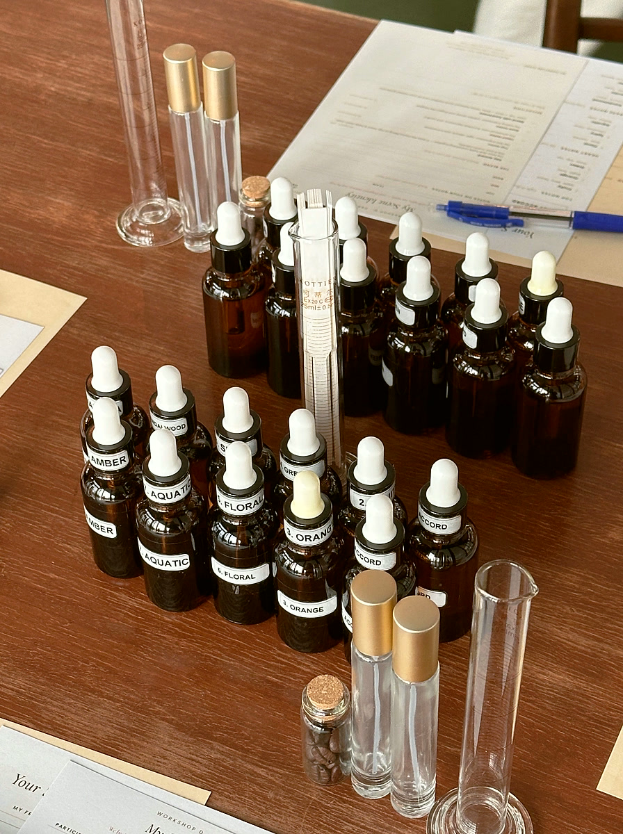 The workshop bench laid out for a session: amber dropper bottles labelled by accord, gold-capped vials, measuring cylinders, and participant worksheets